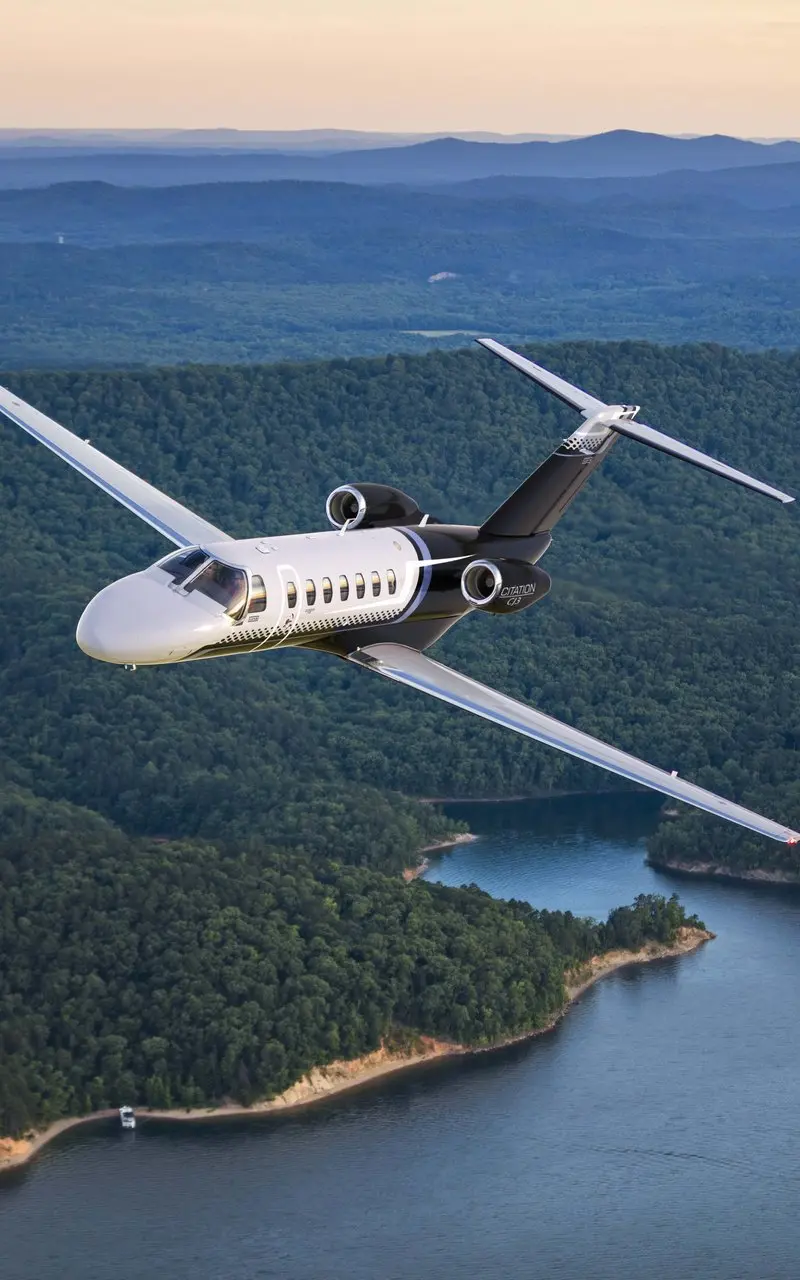Cessna Citation CJ3 — In-flight over scenic landscape The sleek Cessna Citation CJ3 private jet soars gracefully over a lush green forested landscape with a winding blue lake below.