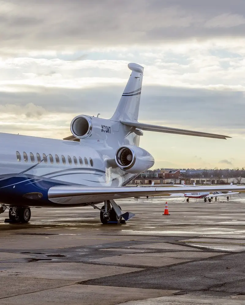 A sleek white and blue Dassault Falcon 7X private jet is parked on a wet tarmac under a cloudy sky, with other aircraft in the background.