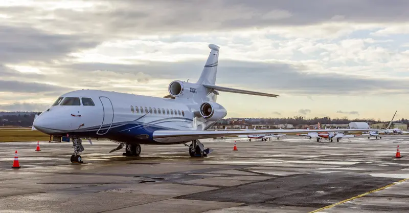 Dassault Falcon 7X — Parked on tarmac A sleek white and blue Dassault Falcon 7X private jet is parked on a wet tarmac under a cloudy sky, with other aircraft in the background.