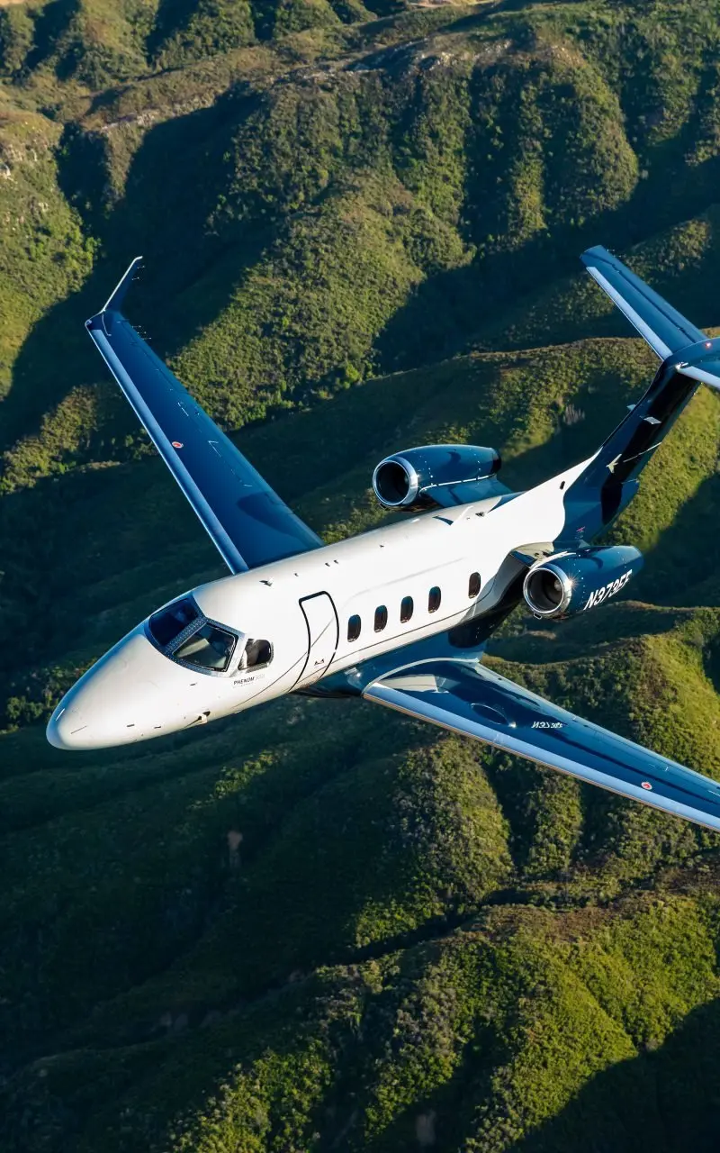 Embraer Phenom 300E — Flying over mountains The Embraer Phenom 300E light jet soars gracefully above lush green mountain ranges near Barcelona.
