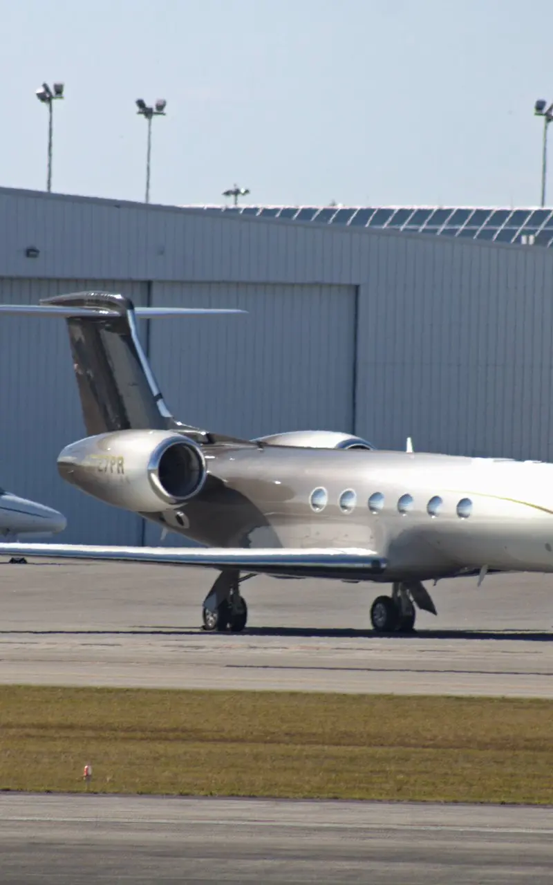 Gulfstream GV — Parked on tarmac, side view A sleek Gulfstream GV private jet is parked on the tarmac, seen from its side, with another smaller jet and a hangar in the background at an airport.