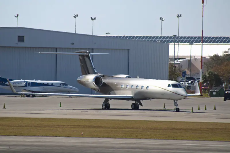 Gulfstream GV — Parked on tarmac, side view A sleek Gulfstream GV private jet is parked on the tarmac, seen from its side, with another smaller jet and a hangar in the background at an airport.