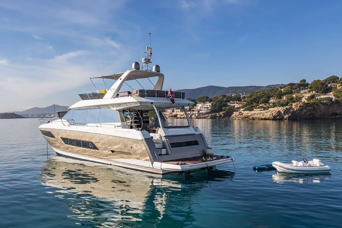 Prestige 680 Fly — Anchored in Marbella bay. The Prestige 680 Fly motor yacht is seen from its stern, anchored in a sunny bay near Marbella, with a tender floating nearby.